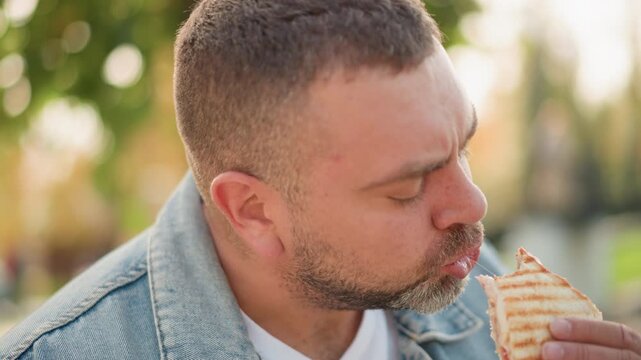 Close up of man eating grilled sandwich while wearing denim jacket and white t shirt. Sandwich held in hand partially eaten with grill marks visible. Outdoors setting with blurred greenery