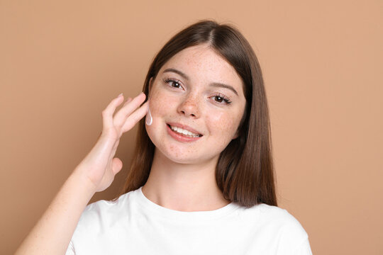 Beautiful teenage girl with freckles applying cream onto her face against light brown background