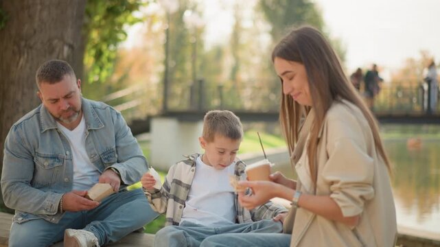 Family enjoying picnic outdoors with food and drinks in park setting man sits on bench eating sandwich while boy interacts nearby woman stands offering drink to child