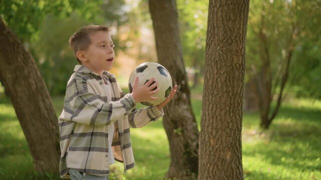 Young boy holding soccer ball in outdoor park setting walking and standing near trees with green grass and autumn foliage. child wears casual clothing including plaid shirt over t shirt
