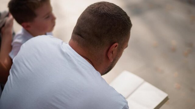 Close up of man and young boy reading together outdoors. Book held open on lap soft natural light calm expression focus on shared activity. Background blurred with neutral tones suggesting