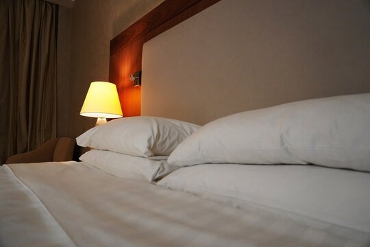 Close-up of hotel bed with white linens and pillows, warm bedside lamp creating cozy atmosphere.