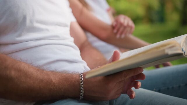 Close up of two people sitting together one holding open book. scene shows hands and torsos with focus on act of reading set against soft outdoor greenery in natural light. Details include
