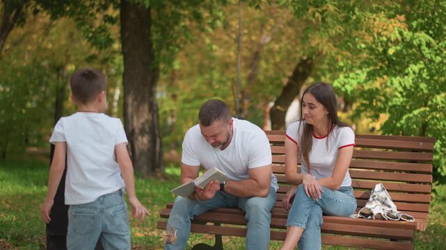 Family enjoying peaceful moment in park. man reads from book while sitting on bench accompanied by woman and their young son who is playing with leaves nearby. scene features green trees