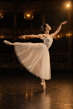 Elegant ballerina in classical white tutu performing arabesque on pointe on a dark vintage theater stage with warm chandeliers