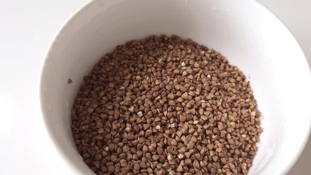 Pouring dry raw buckwheat grains into white ceramic bowl for cooking, close up view