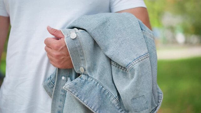 Close up of man holding folded denim jacket over white t shirt in outdoor setting. Jacket shows visible stitching and button details with soft natural light highlighting fabric texture