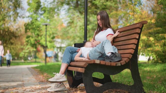 Close up of young woman sitting on park bench with child both relaxed in casual clothing. Woman wears white t shirt and blue jeans background shows green trees path and distant people