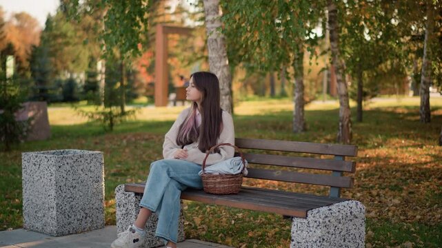 Young woman sits on wooden bench in outdoor park setting wearing casual clothing and holding her hands. wicker basket with folded fabric rests beside her as woman looks around serene