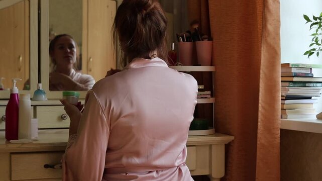 Woman sitting at a vanity table holding an open cosmetic cream jar and applying skincare product in a cozy bedroom setting, beauty and self-care routine concept
