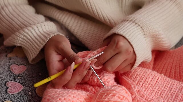 Close-up of a woman crocheting a peach-colored garment with a yellow hook while wearing a cozy beige sweater, handicraft and hobby concept