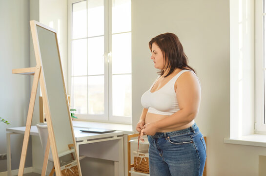 Side view of fat young overweight plump woman measure the waist with measuring tape looking in the mirror standing in living room at home. Slimming, obesity and weight control concept.