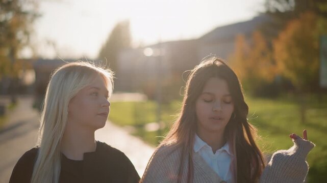 Two young women stand outdoors in park setting during golden hour looking ahead with calm expressions. One wears dark top while other has on light knitted sweater over collared shirt their
