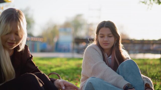 Two young women sit on grass in park engaged in conversation. One wears cream sweater and jeans other is dressed darker with long blonde hair. scene features soft sunlight greenery trees
