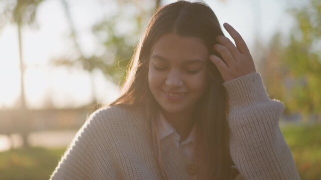 Close up of young woman with long hair wearing knitted sweater and collared shirt sitting outdoors in soft sunlight. Her expression is calm as woman looks around occasionally adjusting her