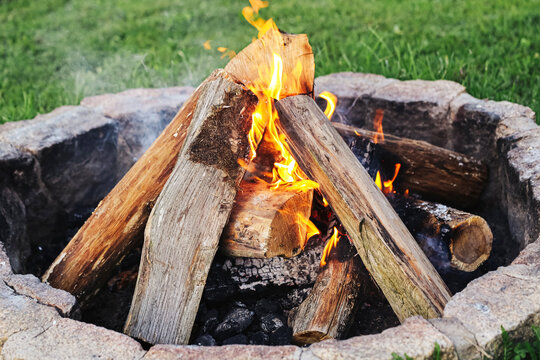 Burning firewood inside circular stone fire pit on green lawn. Orange flames dry logs outdoor campfire garden fireplace rustic heat camping recreation leisure and natural fire background.