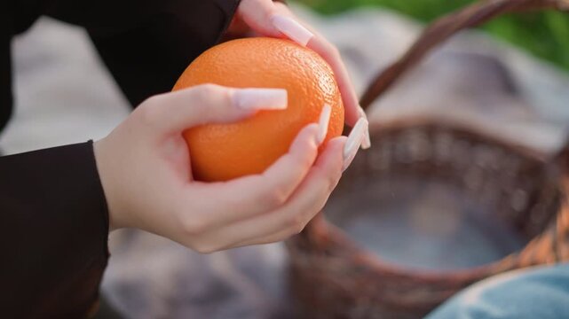 Close up of woman reaching into wicker basket to pick orange examining it closely with long nails. spiral bound notebook lies nearby on fabric suggesting outdoor leisure time. Soft natural