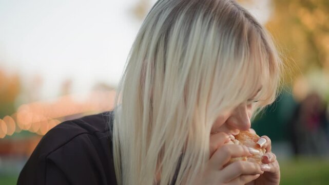 Close up of young woman eating orange in soft natural light. Woman holds fruit with manicured nails focusing on her hands and mouth as woman takes bites. background features blurred autumn