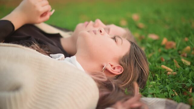 Two women lie close together on grass one with hand resting near other arm. Soft lighting highlights their relaxed expressions and casual clothing details like beige sweater and black top