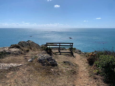 A peaceful, wide-angle shot of a lone green wooden bench situated on a rugged clifftop along the south coast of Guernsey. The bench is perfectly positioned to offer a spectacular panoramic view