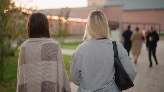 Two women walk away from camera on paved path one wearing patterned shawl and other in gray sweatshirt with black bag. Blurred figures are visible walking ahead near buildings under warm