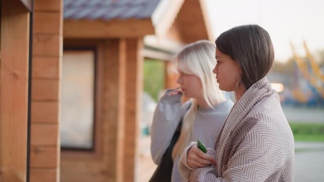Two women stand outdoors near wooden structure engaged in conversation. One holds object while other listens attentively both dressed casually with one wearing patterned wrap and holding