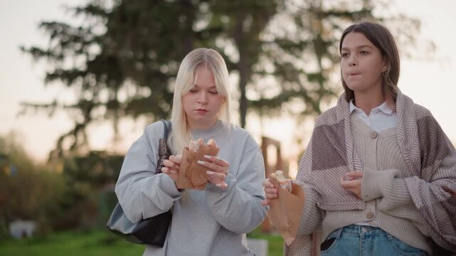 Two young women enjoy eating pastries outdoors in park setting. One wears gray sweatshirt and carries black bag other sports patterned cardigan over white shirt with blue jeans. Soft natural