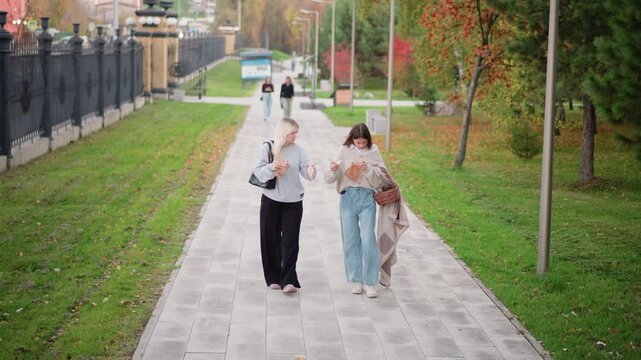 Two young women walk together on paved path in outdoor park each holding food items and carrying bags. One wears black pants with gray top while other has light blue jeans and beige sweater