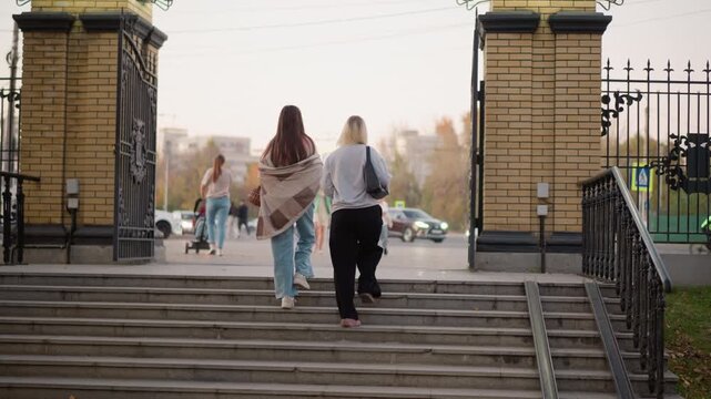 Two women walk up stone steps toward open gate with brick pillars and wrought iron fencing. One carries patterned shawl other has shoulder bag both wear casual clothing suitable for cool