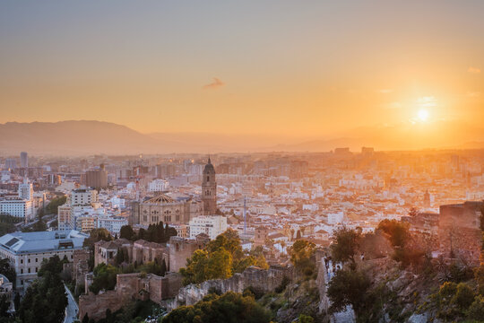 Elevated sunset view of Malaga Cathedral in Spain, rising above dense old town rooftops with Alcazaba walls and distant mountains beneath a glowing orange sky, featuring malaga old town and skyline
