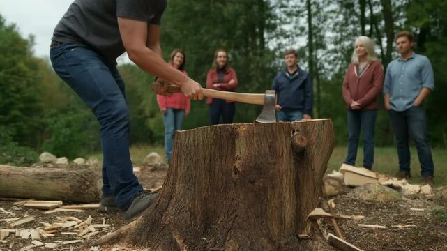 Man swings an axe into a large tree stump while five adults stand nearby watching in a wooded outdoor clearing surrounded by logs, wood chips, rocks, and green trees
