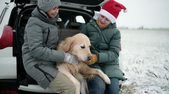 Mother And Daughter Happily Resting With Dog In A Car Trunk Wearing Santa Hats On A Winter Road Stop