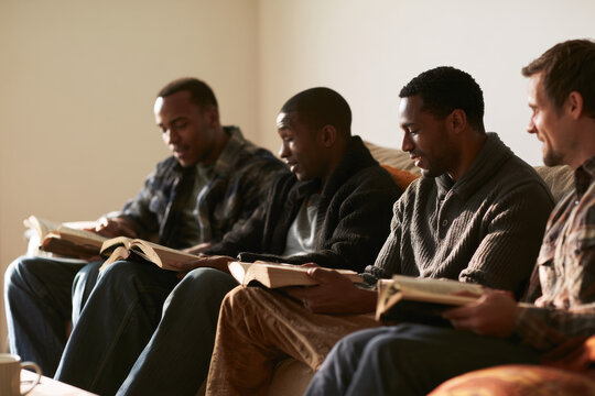 A group of men in their twenties sits on an ordinary living room couch with plain books open, in quiet small-group discussion. Bright, domestic, wide copy space. Bible study Christian prayer meeting