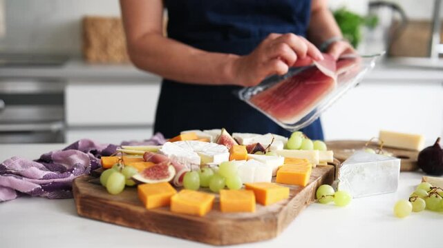 Woman Prepares A Cheese Plate In The Kitchen And Adds Jamon On A Cheese Board