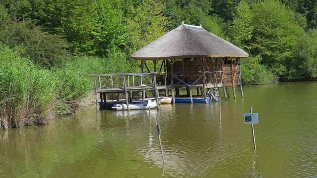 Rustic wooden lakeside shelter on stilts with small boats surrounded by reeds and forest