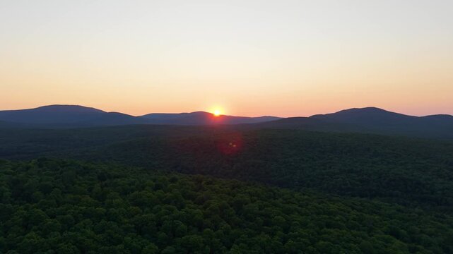 Northern Vermont sunset aerial video over dense forest canopy with golden sun dipping behind silhouette mountain ridges under a warm sky.