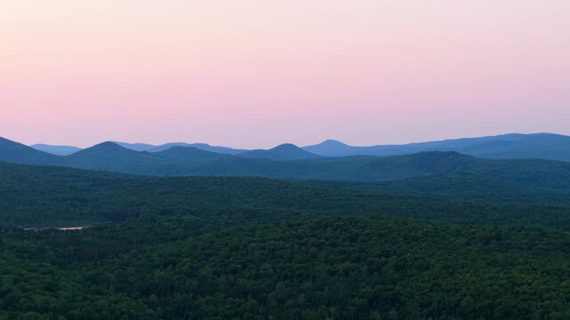 Layered blue mountain ridges and dense green forest canopy under a soft pink pastel sky at sunset in Northern Vermont.