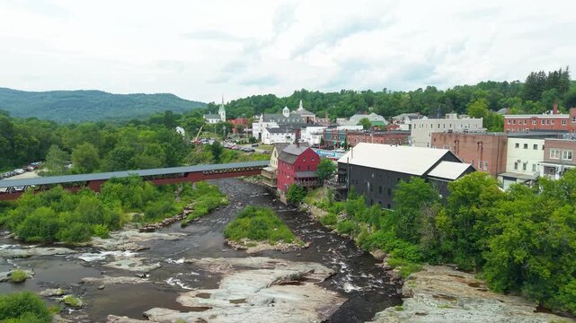 Littleton, New Hampshire, high aerial drone video over the Ammonoosuc River, showing the historic covered bridge, red grist mill, and downtown village architecture set against forested green hills.