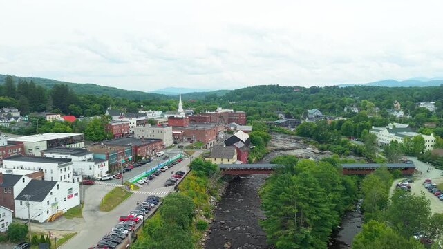 Littleton, New Hampshire, high aerial drone video over the Ammonoosuc River, showing the historic covered bridge, red grist mill, and downtown village architecture set against forested green hills.