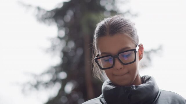 Close up of young woman wearing black glasses and dark jacket standing outdoors with trees in soft focus background. Her hair is tied back fine details include eyeglasses reflection