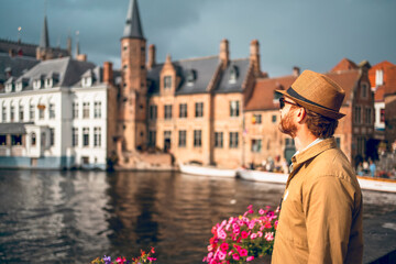A man stands by a canal in Bruges looking at traditional medieval brick and gabled houses, historic structures with towers, pointed spires, and traditional Flemish architecture. © Agnieszka Gaul