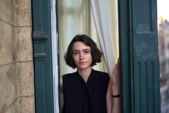 Woman Standing by a Window Holding Curtains in a City