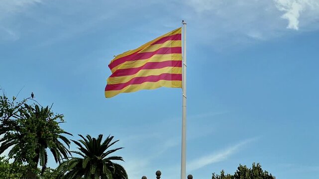 August 2026. Barcelona, Spain. Catalan flag waving on a tall flagpole above historic architecture and green trees on a sunny summer day