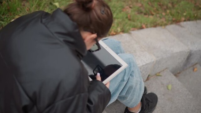 Close up of young woman sitting on stone steps outdoors focused on using her tablet. Woman wears casual clothing including blue jeans and black shoes with anklet surrounded by grassy area