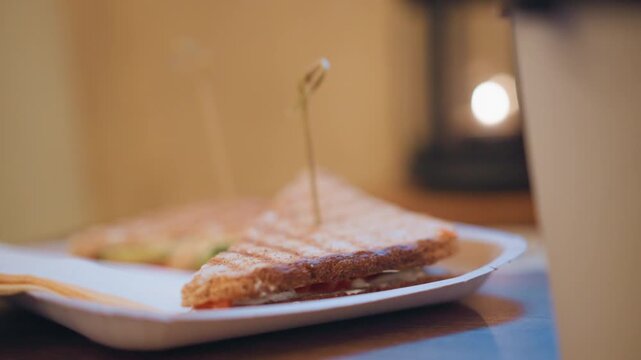 Close up of toasted sandwich on white paper plate with toothpick showing bread texture and filling layers. Background softly blurred to emphasize food item. Warm lighting creates cozy