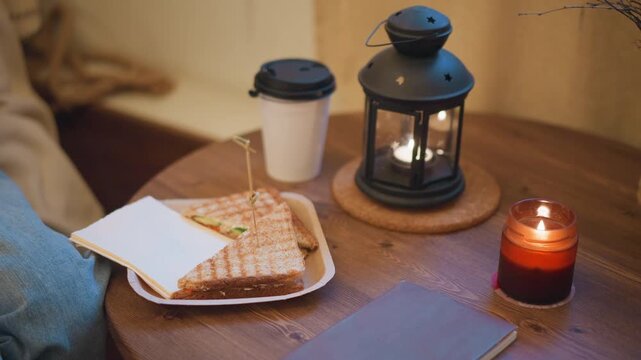 Close up of person lap with blue jeans featuring grilled sandwich on paper plate held by hand white coffee cup and black lid nearby lit candle in glass jar at table corner wooden round table