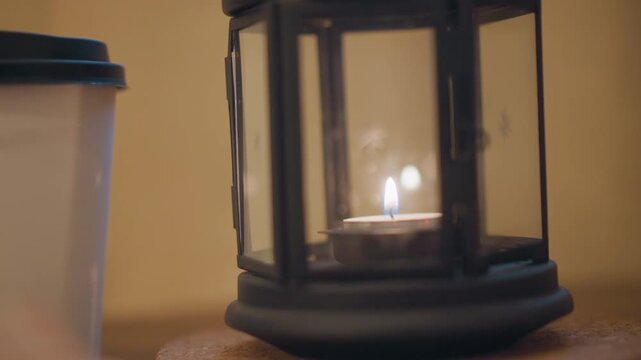 Close up of lit candle in glass lantern next to white cup with black lid on wooden surface. Candle flame flickers gently casting warm light and soft shadows around objects against plain