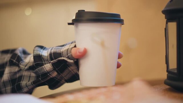 Close up of hand in plaid sleeve reaching for and holding white disposable coffee cup with black lid. scene includes soft lighting bokeh background lights warm indoor setting with wooden