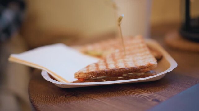Close up of toasted sandwich on white paper plate with toothpick accompanied by open notebook. scene features soft lighting and warm indoor environment details like wooden table and blurred