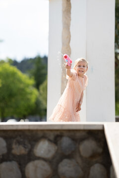 Young girl in a pink dress playfully sprays water with a bright squirt gun while hiding behind a white pillar in a sunny park with soft focus greenery in the background providing depth